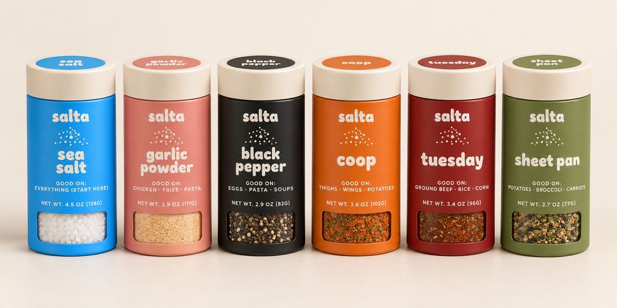 Six salta seasoning jars in a row on a counter — sea salt, garlic powder, black pepper, coop, tuesday, and sheet pan — each a different color with a cream lid and a clear fill window.